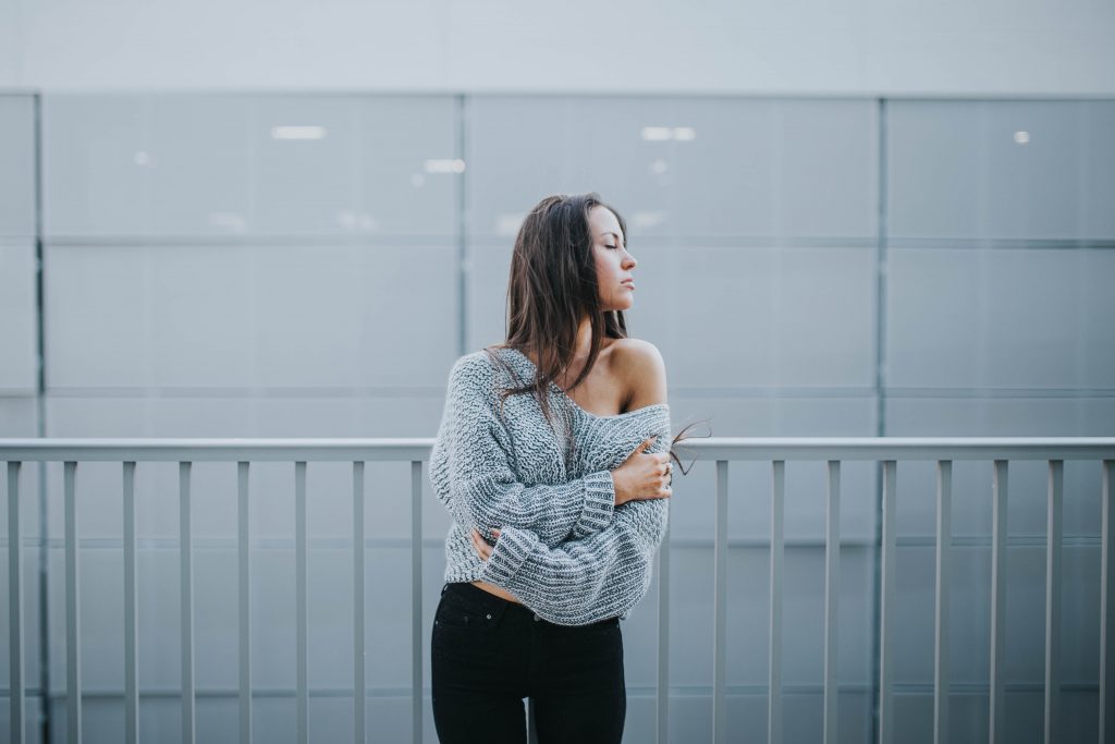 femme brune portant un pull gris laissant apparaitre une épaule et un pantalon noir