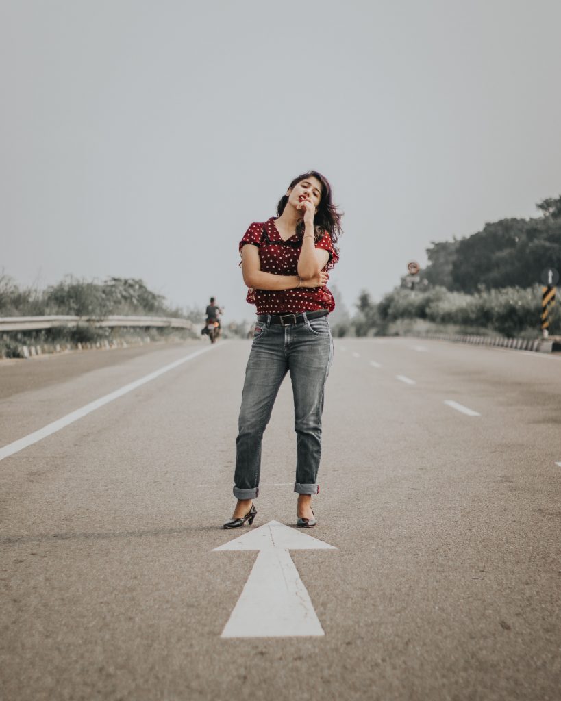 femme se tenant debout au milieu d'une route portant un haut rouge un jean gris et des chaussures à talons